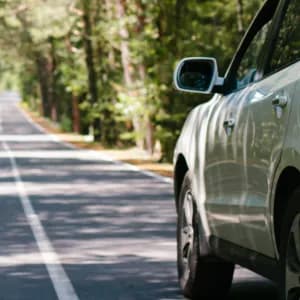 En vit bil kör längs en asfalterad väg kantad av höga, gröna träd. Solljus filtrerar genom löven och kastar fläckiga skuggor på vägen och fordonet.